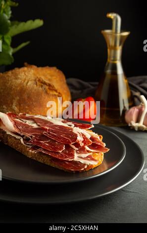 Pane tostato con prosciutto iberico su piastre di nero e sfondo rustico, in background il pane artigianale, pomodoro fresco e olio di oliva Foto Stock