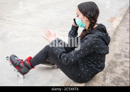 Donna in maschera protettiva sul pattinaggio a rotelle pausa, seduta sulla strada e utilizzando il telefono cellulare durante l'epidemia di coronavirus pandemic. Urban Girl che parla al telefono, indossando pattini a rotelle . Foto Stock