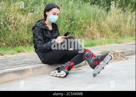 Donna in maschera protettiva, in pausa pattinaggio a rotelle, seduta sulla strada e utilizzando il telefono cellulare durante l'epidemia di coronavirus pandemic. Urban Girl con telefono, con pattini a rotelle . Foto Stock