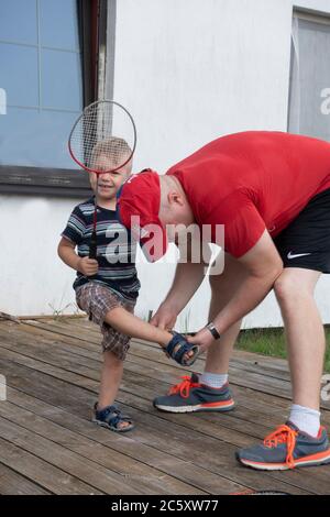 Padre che aiuta il figlio a indossare i sandali e prepararsi a giocare a badminton. Zawady Gmina Rzeczyca Polonia Foto Stock