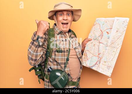 Uomo anziano con capelli grigi che indossa zaino hiker tenendo la mappa che punta il pollice verso il lato sorridente felice con la bocca aperta Foto Stock