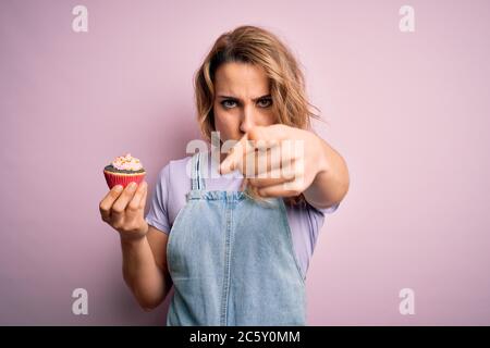 Giovane bella donna bionda eatimg cioccolato cupcake su isolato sfondo rosa puntando con il dito alla macchina fotografica e a voi, segno della mano, positivo Foto Stock