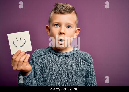 Giovane ragazzino caucasico che mostra faccia sorridente su una nota di carta come messaggio felice spaventato in shock con una faccia sorpresa, impaurito ed eccitato con espe di paura Foto Stock