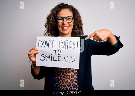 Donna di mezza età che tiene bandiera con non dimenticare il messaggio di sorriso su sfondo bianco molto felice che punta con mano e dito Foto Stock