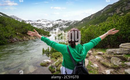 Red head ragazza felice alzò le mani e guarda la montagna. Una donna in felpa verde con zaino si alza con la schiena alla fotocamera Foto Stock