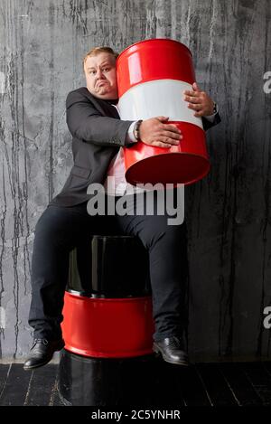 avido ragazzo grasso sul punto di rovinare la sua azienda petrolifera, l'uomo siede hugging rosso barile di petrolio. industria di produzione di petrolio, concetto di mercato globale Foto Stock