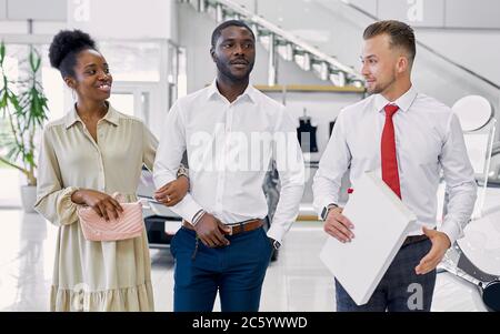 uomo di vendita caucasico sicuro che vende le automobili alla concessionaria di automobile, sta presentando le offerte speciali ai clienti afroamericani, hanno conversazione Foto Stock