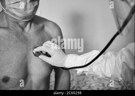 Immagine in bianco e nero. Immagine ravvicinata della mano del medico in tuta protettiva DPI che esamina il paziente anziano di sesso maschile a casa utilizzando un fonendoscopio, controllando il petto. Concetto medico, sanitario Foto Stock