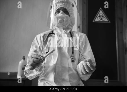 Snapshot bianco e nero di personale medico che tiene il kit di raccolta con tampone COVID-19, indossando tuta protettiva bianca PPE, maschera facciale, guanti, provetta per il prelievo di campioni paziente OP NP Foto Stock