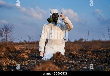 Ambientalista maschile in tuta e maschera a gas che versa il terreno dalla sua mano mentre fa lavori di ricerca su territorio bruciato, analizzando le condizioni del suolo. Concetto di ecologia, ricerca e terra bruciata. Foto Stock