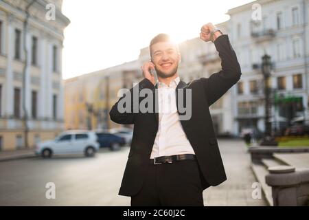 Giovane uomo sorridente con giacca nera e camicia bianca che alza felicemente la mano mentre parla sul cellulare con una splendida vista sulla città sullo sfondo Foto Stock