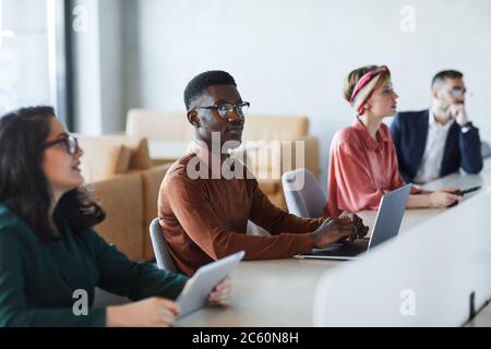 Gruppo multietnico di giovani uomini d'affari seduti al tavolo nella sala conferenze durante il seminario di formazione, concentrarsi su un giovane afro-americano che guarda un Foto Stock