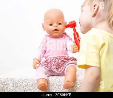 La bambina ispeziona le orecchie della bambola su uno sfondo bianco. Il concetto di otolaringologia pediatrica otite media delle orecchie e test di zolfo in t Foto Stock