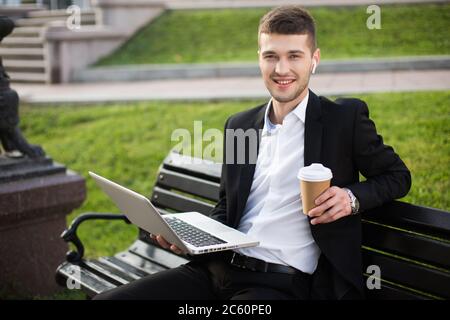 Giovane uomo d'affari bello in giacca nera classica e camicia bianca con auricolari wireless e caffè da andare seduto sulla panca con il computer portatile mentre Foto Stock