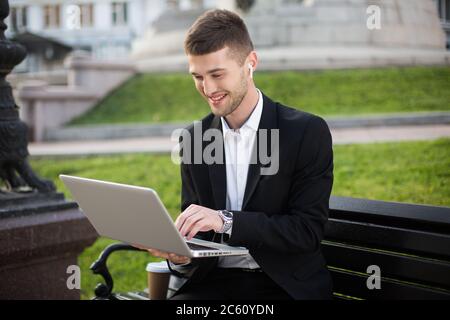 Giovane uomo d'affari sorridente in classica giacca nera e camicia bianca con auricolari wireless seduti sulla panca che lavorano felicemente con il computer portatile mentre si spende Foto Stock