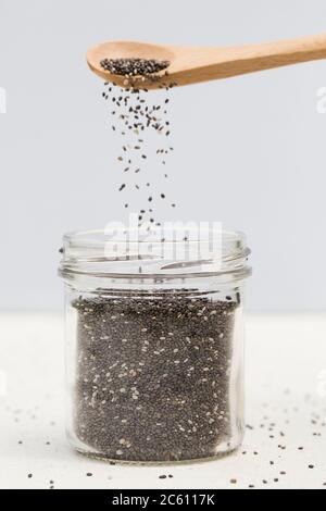 i semi organici di chia vengono versati dal cucchiaio di legno in un vaso di vetro su sfondo grigio. Concetto di dieta sana. Cibo eccellente, fuoco selettivo Foto Stock