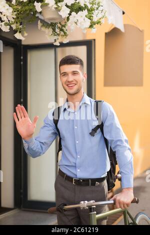 Giovane uomo sorridente in camicia blu con auricolari wireless e zaino in piedi con bicicletta mentre si fa un'agnello all'aperto Foto Stock