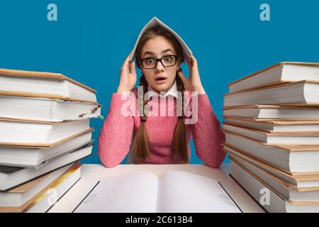 La ragazza adolescente spaventata e scioccata si siede a tavola con pile di libri e copre la testa con il notebook Foto Stock