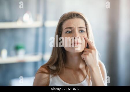 Problema trattamento della pelle. Ragazza sconvolgente con pimple sulla guancia vicino a specchio all'interno Foto Stock