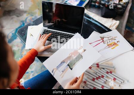 Vista dall'alto della giovane donna seduta al tavolo lavorando su un computer portatile con illustrazioni di moda vicino in officina Foto Stock