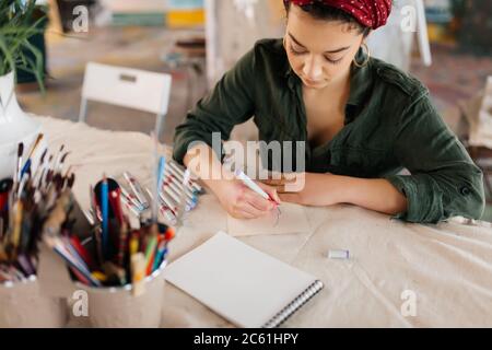 Giovane bella signora con capelli ricci scuri seduta al tavolo disegni disegni sognanti mentre si passa il tempo in accogliente laboratorio da solo Foto Stock