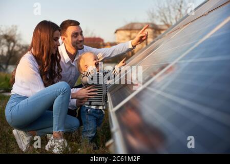 Vista laterale di una giovane famiglia moderna con un bambino familiarizzato con pannello solare in una giornata di sole, verde concetto di energia alternativa Foto Stock