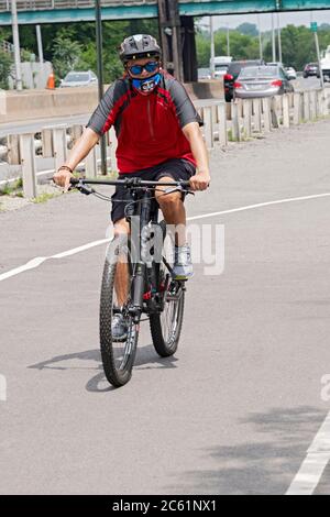 Un uomo americano asiatico che indossa un casco e maschera chirurgica, biciclette su un percorso vicino a thecross Island Parkway. A Bayside, Queens, New York City. Foto Stock