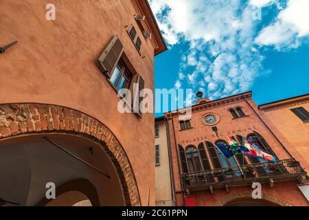 Antica casa storica e municipio con balcone, bandiere e orologio sotto il bel cielo di Alba, Piemonte, Italia settentrionale (vista ad angolo basso). Foto Stock