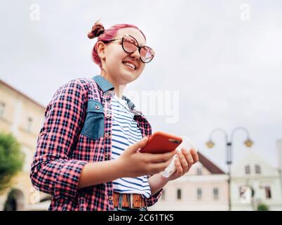 Sorridente bella giovane ragazza moderna ragazza ritratto con straordinaria acconciatura in camicia a scacchi con uno smartphone sottile in mano. Teen moderno Foto Stock