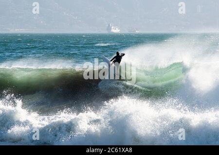 Surfer sulla cresta dell'onda, visto dalla spiaggia di Reñaca, costa centrale del Cile. Sullo sfondo, una nave che arriva al porto di Valparaiso Foto Stock