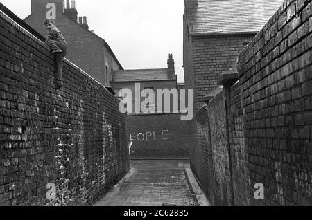 Back to Back, baraccopoli vittoriana che ospita il centro di Belfast, anni '1970, Regno Unito. Un giovane ragazzo si arrampica lungo un alto muro di mattoni in un vicolo di schiena, 1970 HOMER SYKES, Irlanda del Nord Foto Stock