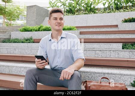 Ritratto di giovane ed eccitato bel uomo d'affari con smartphone in mani seduto sulla panchina e guardando via Foto Stock