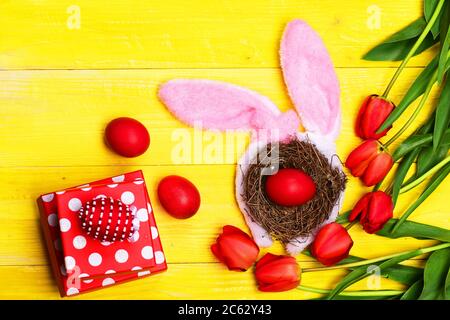 Mazzo di fiori vicino alle uova di Pasqua in nido con orecchie conigliorate poste da scatole presenti. Concetto di simboli di Pasqua. Tulipani in rosa o rosso su sfondo di legno giallo. Composizione delle vacanze primaverili Foto Stock