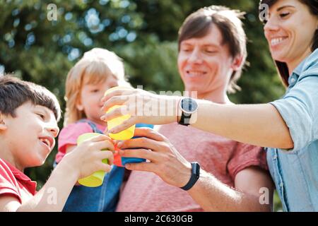 Una famiglia giovane e felice si tostano all'aperto con tazze colorate in un angolo basso, con vista ravvicinata dei volti sorridenti e messa a fuoco sulle mani Foto Stock