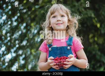 Sorriante giovane bionda ragazza che tiene una ciotola di lamponi freschi maturi succosi in una vista ad angolo basso all'aperto contro le foglie verdi Foto Stock