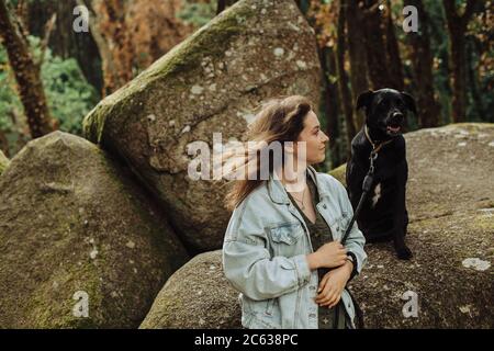 Ragazza che guarda un cane nero su un guinzaglio seduto su roccia nella foresta Foto Stock