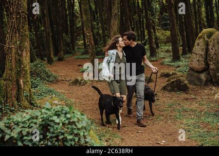 Coppia baciando mentre camminando cani su un guinzaglio giù a. strada in foresta Foto Stock