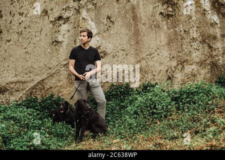 Giovane uomo con due cani neri su un guinzaglio in piedi contro la roccia Foto Stock