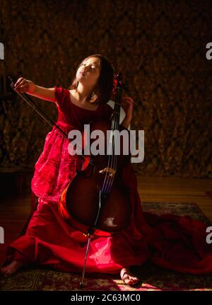 Un piccolo bel bambino in lungo vestito rosso gioca violoncello nella luce del finestrino Foto Stock