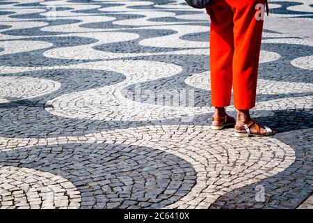 I pantaloni arancioni da donna creano un vivace tocco di colore contro le pietre da pavimentazione con motivi a onda bianchi e neri in Piazza Rossio di Lisbona. Foto Stock