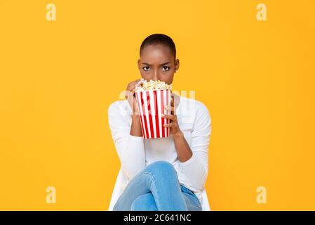 Giovane donna afro-americana spaventata guardando film horror e mangiare popcorn su sfondo giallo isolato Foto Stock