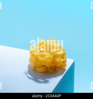Pasta art. Tagliatelle su piedistallo blu pastello su sfondo blu in luce dura. Un tocco creativo di pasta noodle. Design creativo per menu di cucina italiana con spazio per la copia Foto Stock