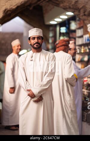 Nizwa / Oman - 15 febbraio 2020: Ritratto di un giovane bell'uomo musulmano Omani che indossa abiti tradizionali bianchi in piedi nel souq coperto di Nizwa con cancello ad arco Foto Stock