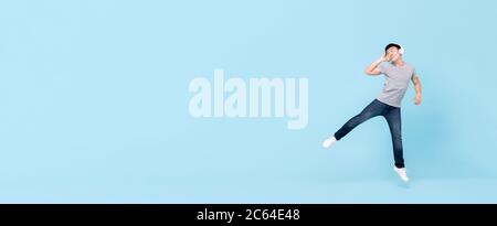 Sorridente giovane asiatico che indossa cuffie ascoltando musica saltando e guardando spazio copia su banner azzurro sfondo isolato Foto Stock