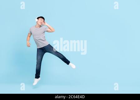 Sorridente giovane asiatico che indossa cuffie ascoltando musica saltando e guardando lo spazio di copia su sfondo azzurro isolato Foto Stock
