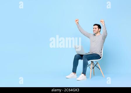 Giovane e ecstatico uomo caucasico vincitore che alza entrambi i pugni con il computer portatile sul grembo contro sfondo azzurro con spazio di copia Foto Stock