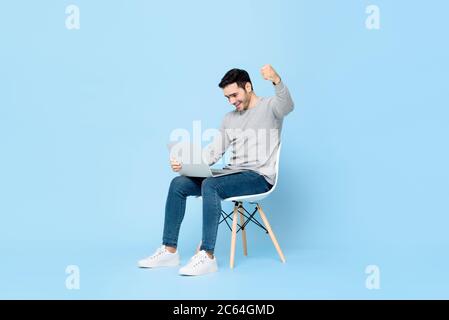 Surpried giovane uomo caucasico guardando il computer portatile e sollevando il suo pugno facendo sì gesto isolato su sfondo azzurro Foto Stock