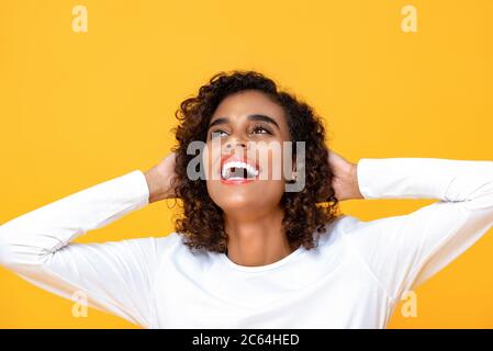 Primo piano ritratto di allegra giovane bella donna afroamericana guardando con entrambe le braccia dietro la testa in uno studio isolato sfondo giallo Foto Stock