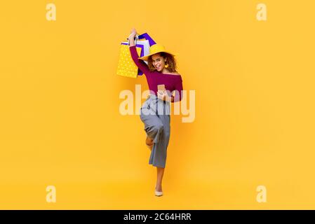 Divertente ritratto a lunghezza intera di felice giovane attraente shopaholic afroamericano donna alzando borse per la spesa e tenendo il telefono cellulare in studio isolato Foto Stock