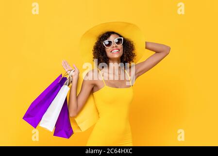 Ritratto estivo di allegra donna afroamericana sorridente che indossa occhiali da sole tenendo il suo cappello e borse per lo shopping in uno sfondo giallo isolato studio Foto Stock